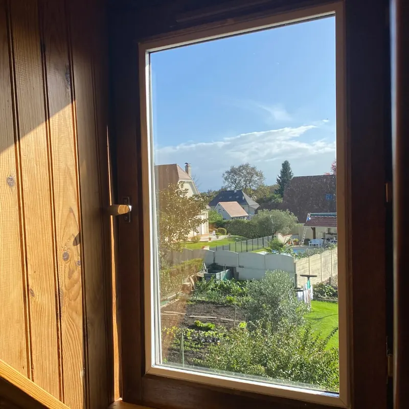 Fenêtre en bois à poignée olive, dans une pièce lambrissée, avec vue sur des jardins et des toits