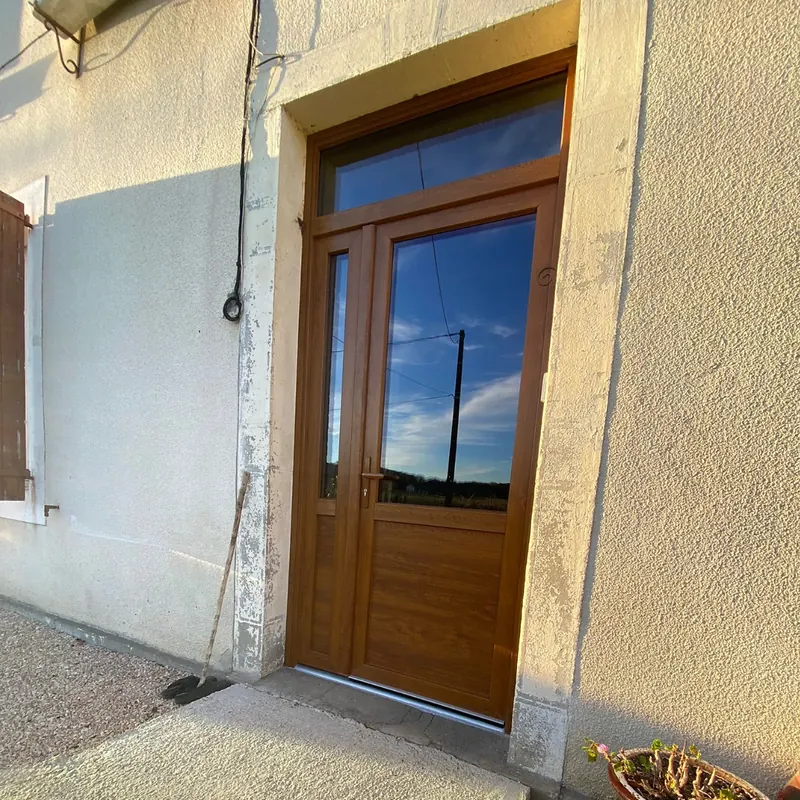 Porte d’entrée à deux vantaux inégaux couleur chêne doré, vitrée, sous une imposte fixe, sur une façade claire