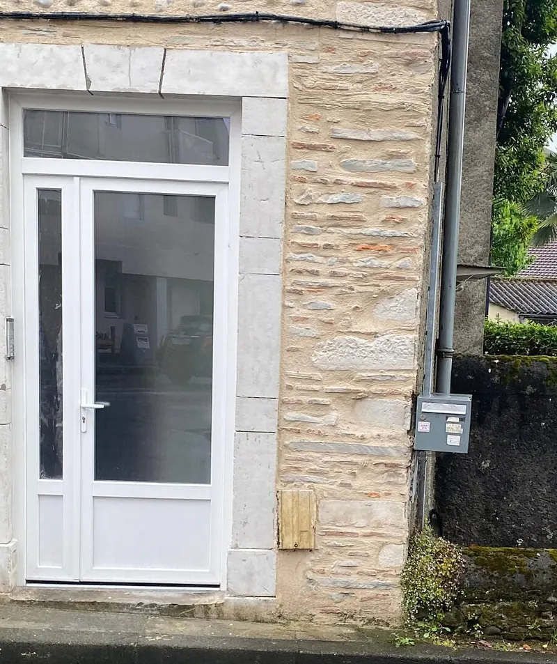 Après : Porte d’entrée PVC blanche vitrée, avec partie latérale étroite et imposte vitrée, posée dans un encadrement en pierre de taille sur une façade en galets et moellons
