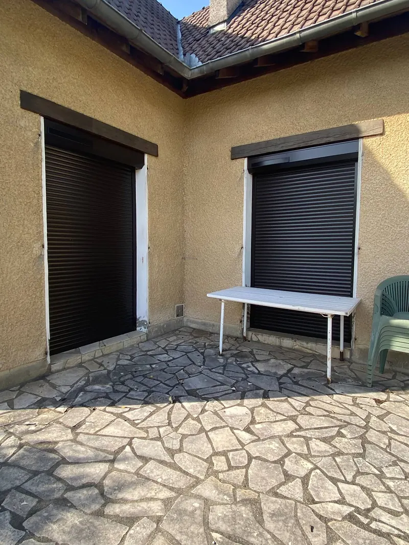 Deux volets roulants noirs fermés sur une porte-fenêtre et une fenêtre, côté terrasse en pierre